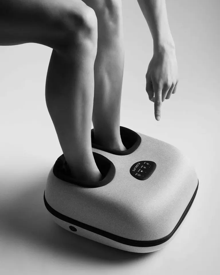 Person using the grey Flowfeet Heat foot massager in a black and white shot, pointing to the top control panel.