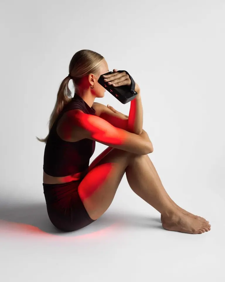 A woman seated in profile uses the Flowlight Panel Go 60 Two Waves for red light therapy, casting a red glow on her skin.