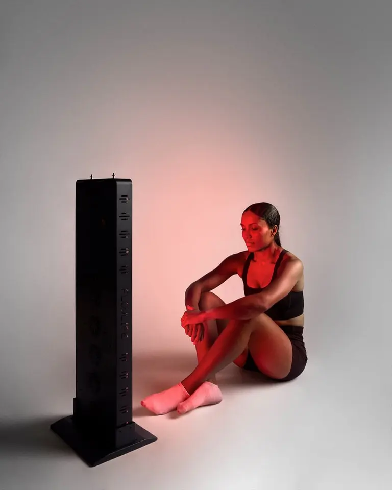 A woman sits in front of the black Flowlight 1500 Seven Waves red light panel during a full-body recovery session.