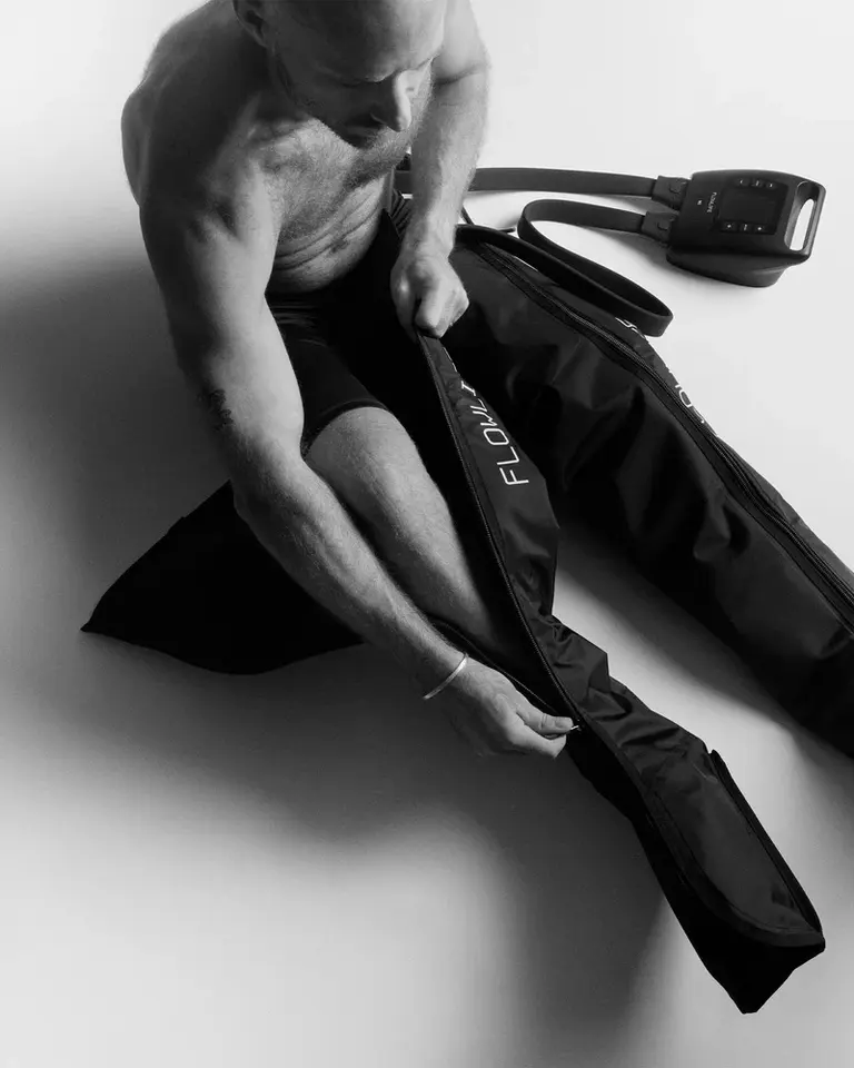 A man zipping up black Flowpression Boots Pro+ for leg recovery in a black and white studio setting.