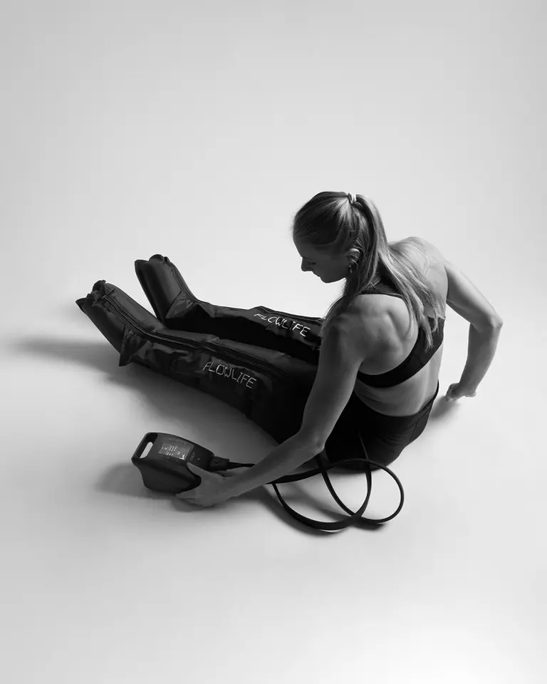 A woman seated on the floor wearing Flowpression Boots Pro+ while adjusting the control unit for leg recovery.