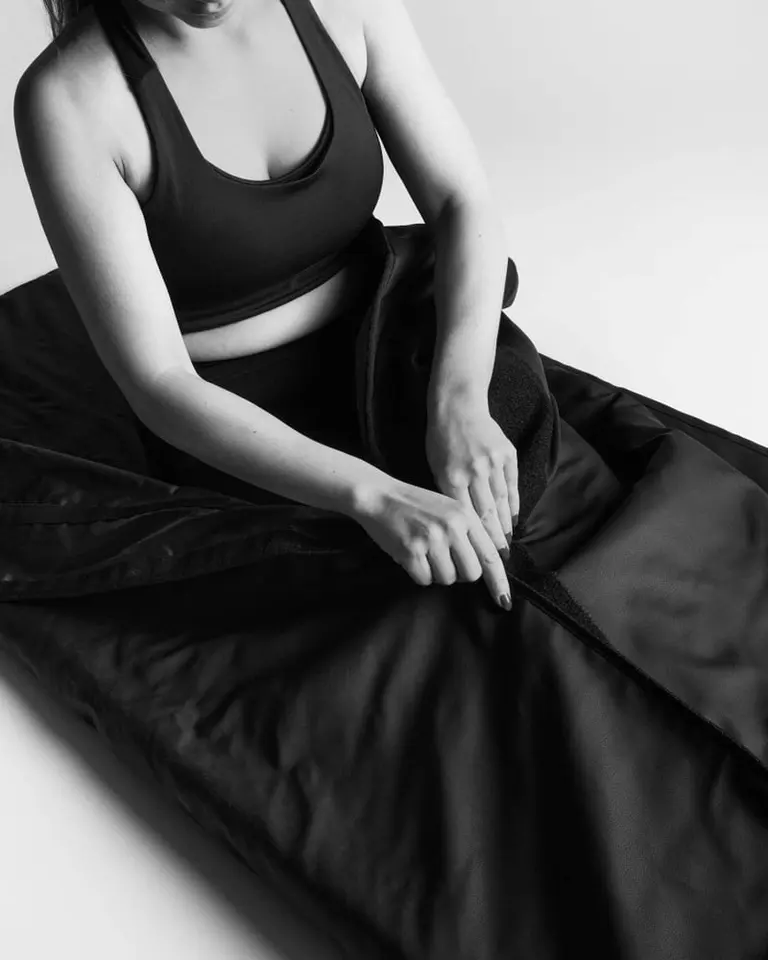 A person fastens the Velcro closure of the black Flowsauna Blanket in a black-and-white studio recovery setting.