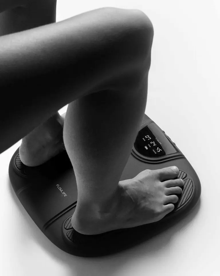 User's bare feet resting on the black Flowtens Feet foot stimulator, showing the integrated LCD display screen.